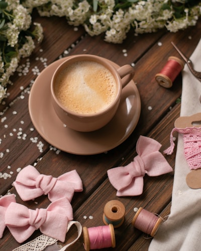 A warm cup of coffee sits on a wooden table surrounded by decorative pink fabric bows, lace, spools of thread, and scattered white flowers, creating a cozy, crafting atmosphere.