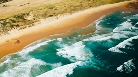 An aerial view of a scenic beach with golden sand stretching alongside vibrant turquoise waters. Gentle waves come ashore, creating a soothing pattern against the coastline. Rolling hills and a grassy landscape are visible in the background, adding a natural, untouched feel to the panorama.