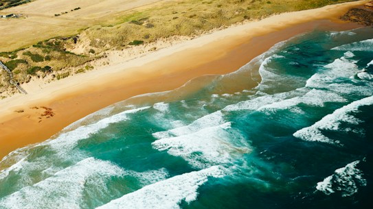 An aerial view of a scenic beach with golden sand stretching alongside vibrant turquoise waters. Gentle waves come ashore, creating a soothing pattern against the coastline. Rolling hills and a grassy landscape are visible in the background, adding a natural, untouched feel to the panorama.