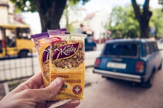 A hand is holding two packs of sunflower seed brittle against a blurred street background. The packages have Cyrillic text and an image of sunflower seeds on the front. In the background, there is a vintage blue car parked on a street and a yellow bus, with leafy trees and buildings in the distance.