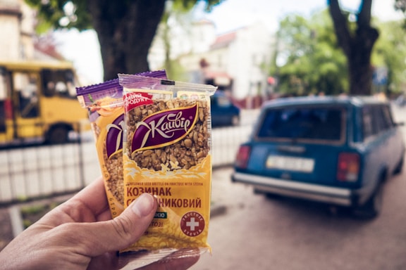A hand is holding two packs of sunflower seed brittle against a blurred street background. The packages have Cyrillic text and an image of sunflower seeds on the front. In the background, there is a vintage blue car parked on a street and a yellow bus, with leafy trees and buildings in the distance.