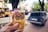A hand is holding two packs of sunflower seed brittle against a blurred street background. The packages have Cyrillic text and an image of sunflower seeds on the front. In the background, there is a vintage blue car parked on a street and a yellow bus, with leafy trees and buildings in the distance.