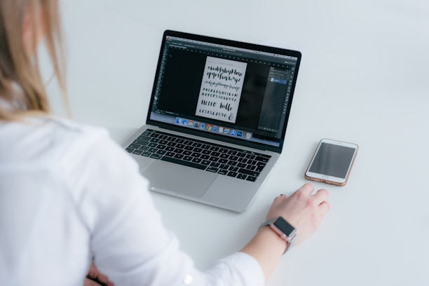 A person wearing a white shirt is using a laptop, with a smartphone placed next to it on a white surface. The laptop screen displays a digital design with various lettering styles.
