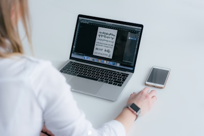 A person wearing a white shirt is using a laptop, with a smartphone placed next to it on a white surface. The laptop screen displays a digital design with various lettering styles.