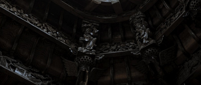 Elaborate wooden carvings and intricate sculptures decorate the ceiling with a dimly lit ambiance. The artwork features detailed figures and patterned designs, showcasing craftsmanship and cultural artistry.