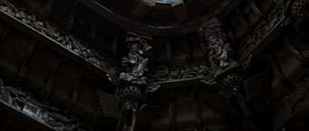 Elaborate wooden carvings and intricate sculptures decorate the ceiling with a dimly lit ambiance. The artwork features detailed figures and patterned designs, showcasing craftsmanship and cultural artistry.