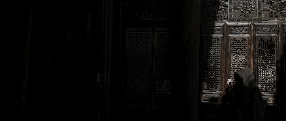 A mysterious figure wearing a hooded cloak stands in front of ornately carved wooden doors with intricate patterns. The setting is dimly lit, creating a shadowy and enigmatic atmosphere. The figure appears to be peering into a small window or gap in the door.