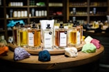 A collection of perfume bottles arranged on a wooden circular display with vibrant colored stones in the foreground. The shelves in the background are filled with various bottles, creating a cozy and inviting setting. Each perfume bottle is labeled with unique names and has a tester tag attached.
