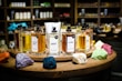 A collection of perfume bottles arranged on a wooden circular display with vibrant colored stones in the foreground. The shelves in the background are filled with various bottles, creating a cozy and inviting setting. Each perfume bottle is labeled with unique names and has a tester tag attached.