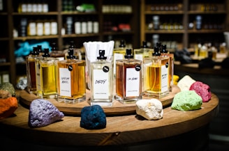 A collection of perfume bottles arranged on a wooden circular display with vibrant colored stones in the foreground. The shelves in the background are filled with various bottles, creating a cozy and inviting setting. Each perfume bottle is labeled with unique names and has a tester tag attached.