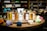 A collection of perfume bottles arranged on a wooden circular display with vibrant colored stones in the foreground. The shelves in the background are filled with various bottles, creating a cozy and inviting setting. Each perfume bottle is labeled with unique names and has a tester tag attached.