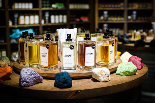 A collection of perfume bottles arranged on a wooden circular display with vibrant colored stones in the foreground. The shelves in the background are filled with various bottles, creating a cozy and inviting setting. Each perfume bottle is labeled with unique names and has a tester tag attached.