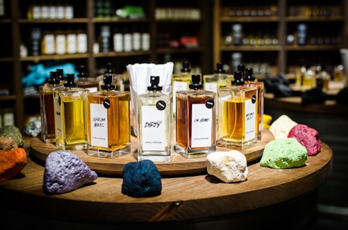 A collection of perfume bottles arranged on a wooden circular display with vibrant colored stones in the foreground. The shelves in the background are filled with various bottles, creating a cozy and inviting setting. Each perfume bottle is labeled with unique names and has a tester tag attached.