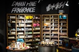A well-organized fragrance shop displays an array of perfume bottles on wooden shelves, with vibrant towels neatly stacked on a few shelves. Two circular tables in the foreground hold a variety of perfume bottles, surrounded by small, colorful fabric pouches and decorative items. Overhead lights illuminate the products, and bold signage on the wall reads 'Damn Fine Fragrance' along with Chinese characters.
