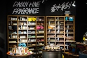 A well-organized fragrance shop displays an array of perfume bottles on wooden shelves, with vibrant towels neatly stacked on a few shelves. Two circular tables in the foreground hold a variety of perfume bottles, surrounded by small, colorful fabric pouches and decorative items. Overhead lights illuminate the products, and bold signage on the wall reads 'Damn Fine Fragrance' along with Chinese characters.