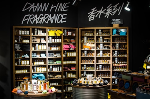 A well-organized fragrance shop displays an array of perfume bottles on wooden shelves, with vibrant towels neatly stacked on a few shelves. Two circular tables in the foreground hold a variety of perfume bottles, surrounded by small, colorful fabric pouches and decorative items. Overhead lights illuminate the products, and bold signage on the wall reads 'Damn Fine Fragrance' along with Chinese characters.