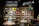 A well-organized fragrance shop displays an array of perfume bottles on wooden shelves, with vibrant towels neatly stacked on a few shelves. Two circular tables in the foreground hold a variety of perfume bottles, surrounded by small, colorful fabric pouches and decorative items. Overhead lights illuminate the products, and bold signage on the wall reads 'Damn Fine Fragrance' along with Chinese characters.