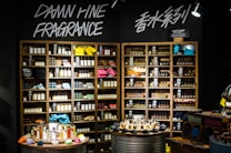 A well-organized fragrance shop displays an array of perfume bottles on wooden shelves, with vibrant towels neatly stacked on a few shelves. Two circular tables in the foreground hold a variety of perfume bottles, surrounded by small, colorful fabric pouches and decorative items. Overhead lights illuminate the products, and bold signage on the wall reads 'Damn Fine Fragrance' along with Chinese characters.