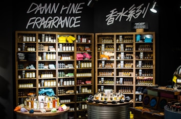 A well-organized fragrance shop displays an array of perfume bottles on wooden shelves, with vibrant towels neatly stacked on a few shelves. Two circular tables in the foreground hold a variety of perfume bottles, surrounded by small, colorful fabric pouches and decorative items. Overhead lights illuminate the products, and bold signage on the wall reads 'Damn Fine Fragrance' along with Chinese characters.