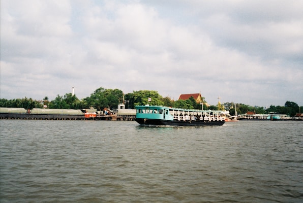A calm river scene with a large turquoise and white boat floating on the water. In the background, there is a line of trees, buildings with triangular roofs, and a cloudy sky. The boat appears to be moving across the river, and there are structures visible along the riverbank.