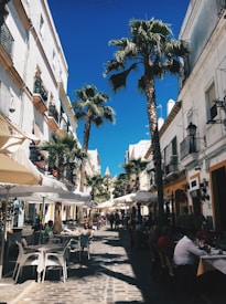 A picturesque cobblestone street flanked by palm trees and lined with cafes. People are seated under white umbrellas, enjoying their time in the sunshine. The architecture consists of colorful buildings with balconies adorned with plants.