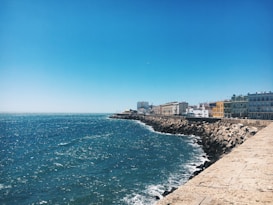 A scenic coastal view with a deep blue sea meeting the sky on the horizon. A paved walkway lined with colorful buildings and a rocky breakwater stretches along the shoreline. The sun shines brightly, creating sparkles on the water surface.