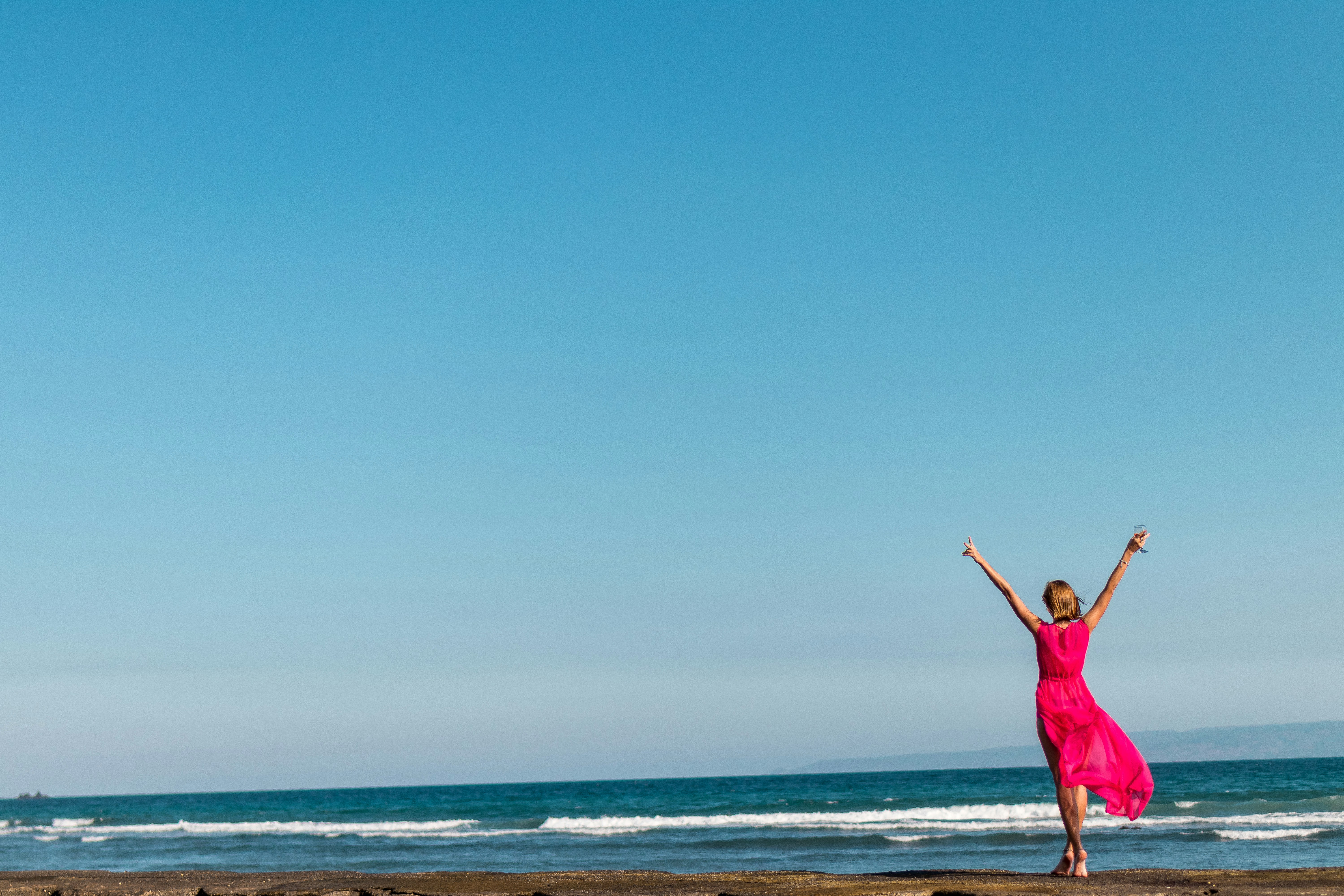 mujer con vestido rosa levantando las manos de pie en la orilla del mar