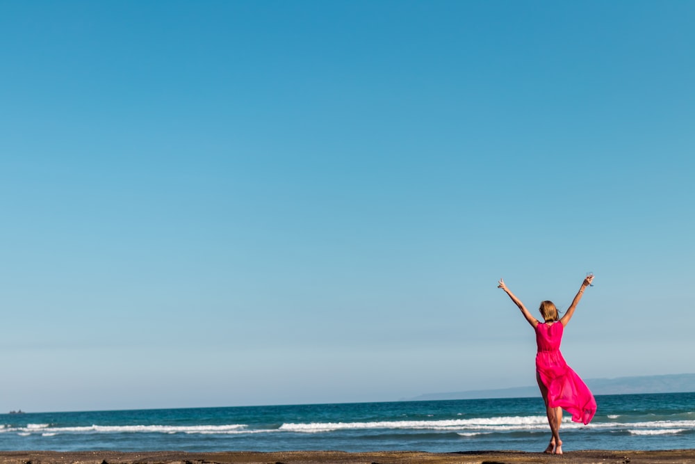 woman wearing pink dress raising her hands standing on sea shore