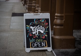 A chalkboard sign placed on a sidewalk, displaying colorful and artistic lettering with the word 'Guava' at the top adorned with decorative swirls and shapes. Below, the vibrant text 'We are OPEN' communicates that the establishment is welcoming customers. The surrounding environment features a brown pillar and paving, creating a cozy and inviting atmosphere.