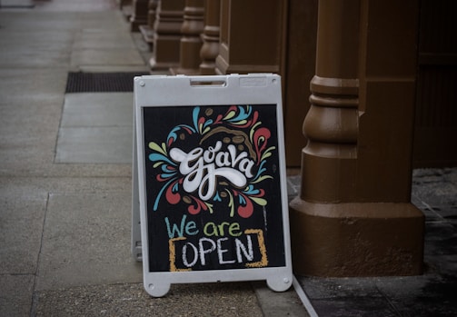 A chalkboard sign placed on a sidewalk, displaying colorful and artistic lettering with the word 'Guava' at the top adorned with decorative swirls and shapes. Below, the vibrant text 'We are OPEN' communicates that the establishment is welcoming customers. The surrounding environment features a brown pillar and paving, creating a cozy and inviting atmosphere.