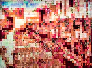 A colorful quilt made up of numerous small square patches of fabric, arranged in a mosaic pattern. The patches display a range of warm colors including reds, pinks, and oranges, interspersed with some greens and whites. The texture of the fabric is visible, with some squares featuring floral and patterned designs.