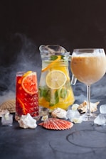 An arrangement of refreshing beverages featuring a pitcher of citrus water with lemon slices and herbs, a tall glass of iced red fruit drink, and a wine glass with a creamy iced coffee. Seashells, ice cubes, and a dark smoky backdrop enhance the aesthetic.