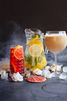 An arrangement of refreshing beverages featuring a pitcher of citrus water with lemon slices and herbs, a tall glass of iced red fruit drink, and a wine glass with a creamy iced coffee. Seashells, ice cubes, and a dark smoky backdrop enhance the aesthetic.