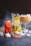 An arrangement of refreshing beverages featuring a pitcher of citrus water with lemon slices and herbs, a tall glass of iced red fruit drink, and a wine glass with a creamy iced coffee. Seashells, ice cubes, and a dark smoky backdrop enhance the aesthetic.