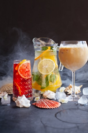 An arrangement of refreshing beverages featuring a pitcher of citrus water with lemon slices and herbs, a tall glass of iced red fruit drink, and a wine glass with a creamy iced coffee. Seashells, ice cubes, and a dark smoky backdrop enhance the aesthetic.