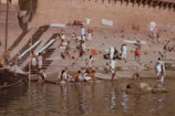 People are gathered on stone steps leading down to a river, possibly bathing or washing clothes. Some are seated or standing, while others are engaged in various activities. There are visible cloths hanging, and the setting appears to be a riverside or a ghat.