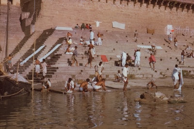 People are gathered on stone steps leading down to a river, possibly bathing or washing clothes. Some are seated or standing, while others are engaged in various activities. There are visible cloths hanging, and the setting appears to be a riverside or a ghat.