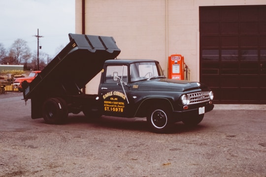A vintage green dump truck is parked with its bed in an elevated position beside a brick garage. The truck has company signage painted on the door in yellow. Beside it is an old red gasoline pump with the logo 'Atlantic.' The surrounding area shows a partially cloudy sky and barren trees in the background, indicating a likely autumn or winter setting.