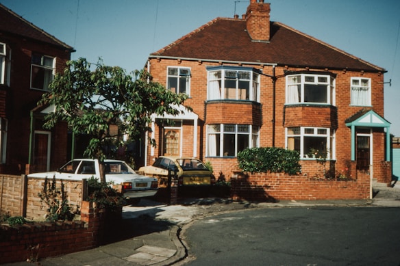 A brick duplex house with bay windows and white frames, surrounded by a small brick wall. The house features a red tiled roof and a green front door. There are parked cars in the driveway on both sides, and a small green tree in the front yard.