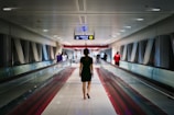 A person walks through an airport or transit corridor lined with glass and metal supports. Others in the distance walk in both directions. Overhead signage guides passengers to a train.