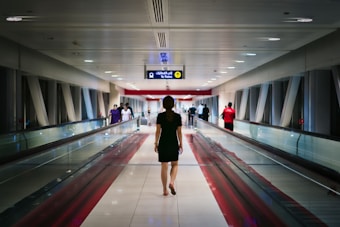 A person walks through an airport or transit corridor lined with glass and metal supports. Others in the distance walk in both directions. Overhead signage guides passengers to a train.