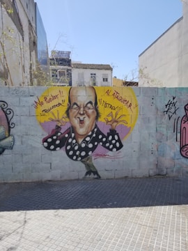 A caricature graffiti of a bald man with an exaggerated facial expression is painted on a brick wall. The man is wearing a black shirt with white polka dots and is shown making a gesture with his hands. Bright yellow and purple colors form a background circle around the character. Spanish phrases '¡No Puedo!! Rectador' and '¡¡Al Ataqueur!! ¡¡Astroz!!' accompany the image. There are other tags and drawings on the wall, and buildings and trees are visible in the background.