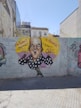 A caricature graffiti of a bald man with an exaggerated facial expression is painted on a brick wall. The man is wearing a black shirt with white polka dots and is shown making a gesture with his hands. Bright yellow and purple colors form a background circle around the character. Spanish phrases '&iexcl;No Puedo!! Rectador' and '&iexcl;&iexcl;Al Ataqueur!! &iexcl;&iexcl;Astroz!!' accompany the image. There are other tags and drawings on the wall, and buildings and trees are visible in the background.