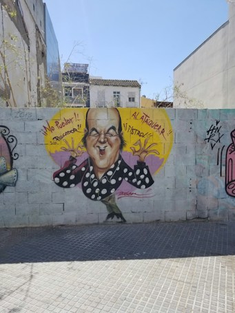 A caricature graffiti of a bald man with an exaggerated facial expression is painted on a brick wall. The man is wearing a black shirt with white polka dots and is shown making a gesture with his hands. Bright yellow and purple colors form a background circle around the character. Spanish phrases '&iexcl;No Puedo!! Rectador' and '&iexcl;&iexcl;Al Ataqueur!! &iexcl;&iexcl;Astroz!!' accompany the image. There are other tags and drawings on the wall, and buildings and trees are visible in the background.