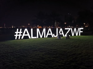 Brightly lit hashtag sign '#ALMAJAZWF' on a grassy field at night, with two people sitting beside it. The background features city lights and silhouettes of palm trees.