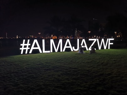 Brightly lit hashtag sign '#ALMAJAZWF' on a grassy field at night, with two people sitting beside it. The background features city lights and silhouettes of palm trees.