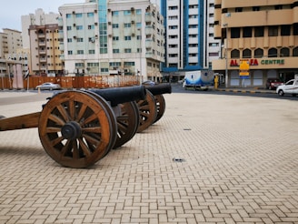 A historical cannon with wooden wheels is placed on a paved open area. Surrounding the cannon are multi-story buildings, including residential apartments and a commercial center named Al Safa Centre. A truck with advertising and several parked cars are visible in the street nearby.