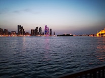 A serene waterfront scene at dusk with a city skyline in the background, featuring a mix of high-rise buildings with vibrant neon lights and a softly illuminated mosque on the right. The sky transitions from blue to a subtle orange near the horizon, reflecting gently on the calm water.