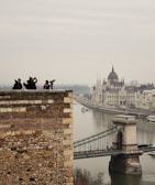 A scenic view of a historic cityscape featuring a prominent river and iconic architecture. In the foreground, several people are standing on a stone wall, some taking photos. The background includes a large, ornate building with spires and a bridge spanning the river.