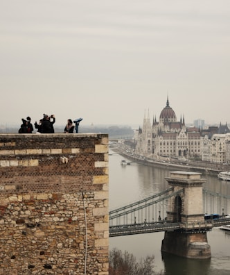 A scenic view of a historic cityscape featuring a prominent river and iconic architecture. In the foreground, several people are standing on a stone wall, some taking photos. The background includes a large, ornate building with spires and a bridge spanning the river.